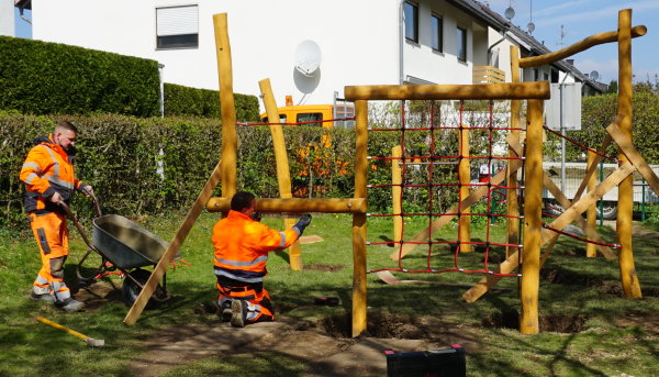 Auf dem Bild sind die Mitarbeiter des Bauhofs zu sehen, die auf einem Spielplatz ein Klettergerüst für Kinder anbringen.