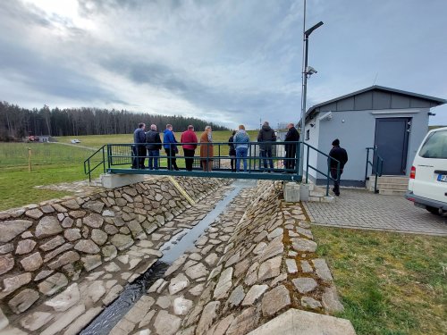 Zu sehen sind die Teilnehmer des Erfahrungsaustauschs. Sie stehen auf einer Metallbrücke über einem Bach, der mit Stein ausgekleidet ist. Zur rechten befindet sich ein Pegelmesshäuschen, vor ihnen ragt das Regenrückhaltebecken Neuwürschnitz auf.