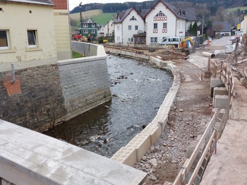 Zu sehen ist ein Bach, der durch eine Ortschaft fließt. Am rechten Ufer wird zwischen den Häusern mit Baggern und schweren Geräten eine hohe Mauer um den Bach gezogen, um ein Übertreten zu verhindern.