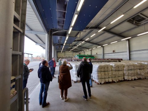 Zu sehen ist eine große Lagerhalle, auf der viele Hochwasserbekämpfungsmittel wie leere Sandsäcke auf Holzpaletten aufgetürmt sind. Zu sehen sind Teilnehmer des Austauschs, die die hoch aufgetürmten Mittel bestaunen.