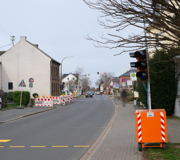 Das Bild zeigt die Essiger Straße in Odendorf auf Höhe des Bahnübergangs. Vor und hinter dem Bahnübergang sieht man die gesperrten Bürgersteige sowie die aufgestellten Bedarfsampeln für Fußgänger.