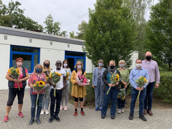 Auf dem Photo sind die elf Mitglieder der Ganztagesbetreuung an der Sekundarschule zu sehen. Sie stehen in zwei Reihen vor einem der Schulgebäude und haben Sonnenblumen in der Hand.