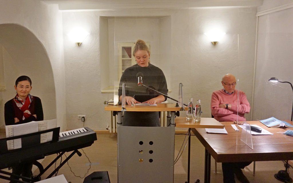 Links an einer Orgel sitzend sieht man Yaein Lee, im Zentrum steht Lara Schmidt an einem Rednerpult und im rechten Teil des Fotos sitzt Armin Schmidt an einem Tisch