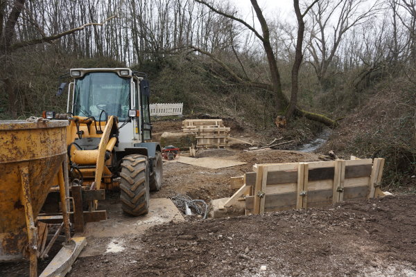 Auf dem Bild sind die Brückenarbeiten an der Brücke Im Klinkert zu sehen. Ein Bagger und verschiedene Holzelemente sind zu sehen.