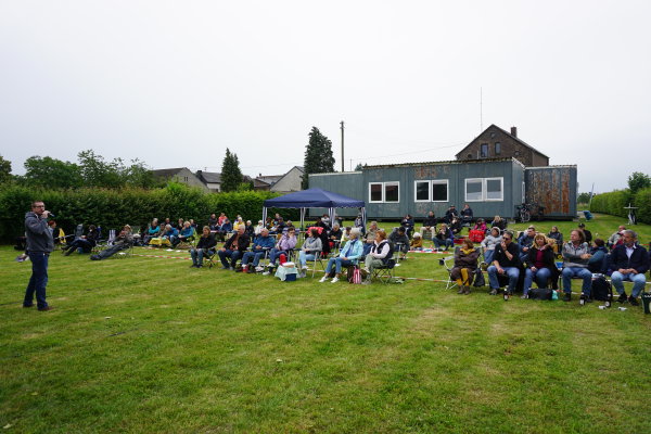 Auf dem Bild ist das Publikum des Picknick-Konzerts zu sehen, das auf einer Wiese hinter der alten Schule in Dünstekoven sitzt. 