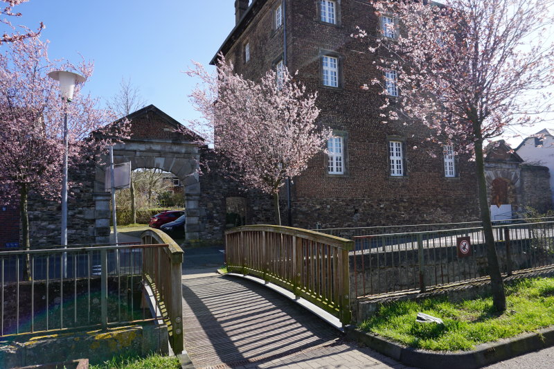 Auf dem Bild ist die Brücke über den Orbach richtung Burg Odendorf zu sehen. Das Bild wurde im Frühling aufgenommen, die Sonne scheint und die Bäume sind in Blüte.