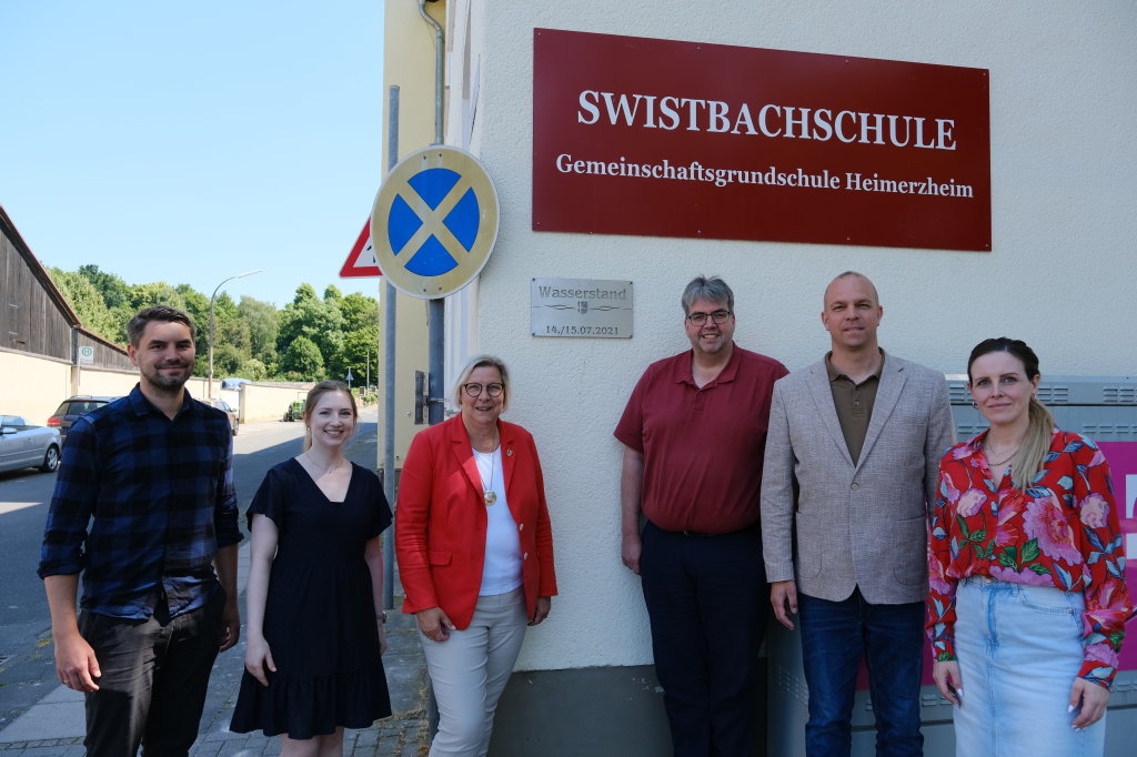 Foto: © Gemeinde Swisttal: v.l.n.r.: Dennis Schönenstein (Stabsstelle Wiederaufbau), Celine Braun (Stabsstellenleiterin Wiederaufbau), Petra Kalkbrenner (Bürgermeisterin), Jörg Timmermann (Ingenieurbüro pbs), Tobias Weingartz (Beigeordneter), Barbara Kolz (Schulleiterin Swistbachschule) Foto vor einer Flutmarke am Gebäude der Swistbachschule in Heimerzheim