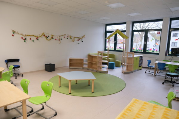 Auf dem Bild sieht man beispielhaft ein Klassenzimmer der Grundschüler. Vorherrschende Farben sind Weiß und Grün; Tische mit Stühlen laden zum Sitzen ein. Im Hintergrund sieht man eine Sitzecke mit Kissen und Sofas.